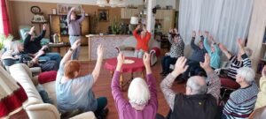 Eine Gruppe von Menschen sitzt auf Stühlen und Couches und streckt die Arme hoch.