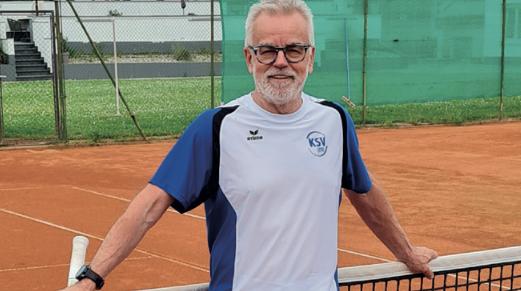 Ein Mann mit grauen Haare, einer schwarzen Brille und einem blau-weißen T-Shirt steht auf einem Tennisplatz und hält sich mit beiden Händen am Netz hinter ihm fest.
