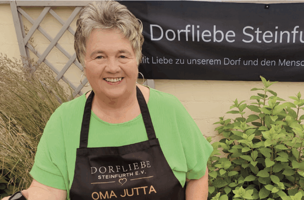 Frau mit kurzen Haaren und schwarzer Schürze auf der "Oma Jutta" steht, die ein Waffeleisen in der Hand hält.
