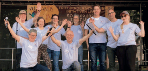 Viele Menschen stehen auf der Bühne mit einem hellblauen T-Shirt und strecken die Hände in die Höhe.