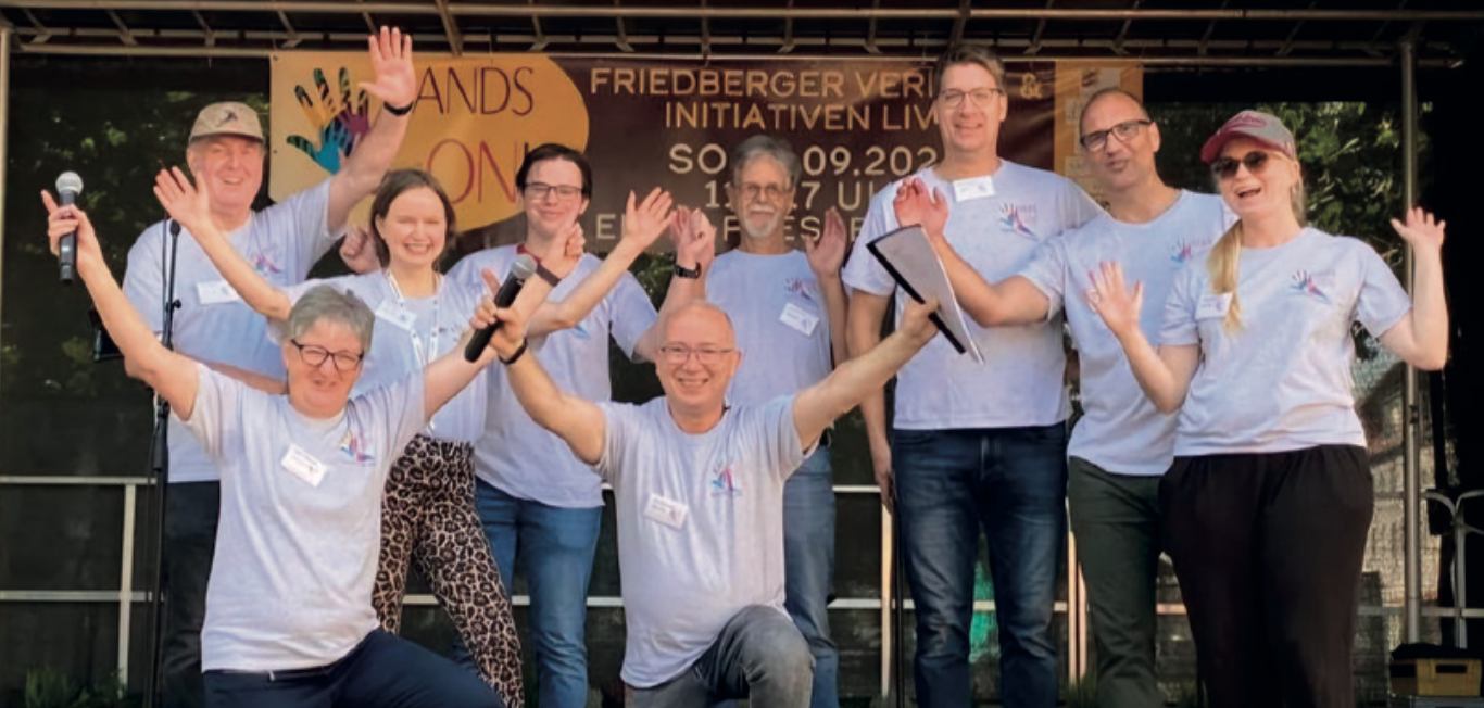 Viele Menschen stehen auf der Bühne mit einem hellblauen T-Shirt und strecken die Hände in die Höhe.