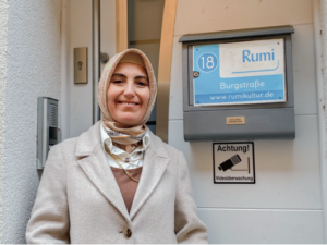 Eine Frau in beigen Klamotten und mit Kopftuch, die vor einem Briefkasten steht.